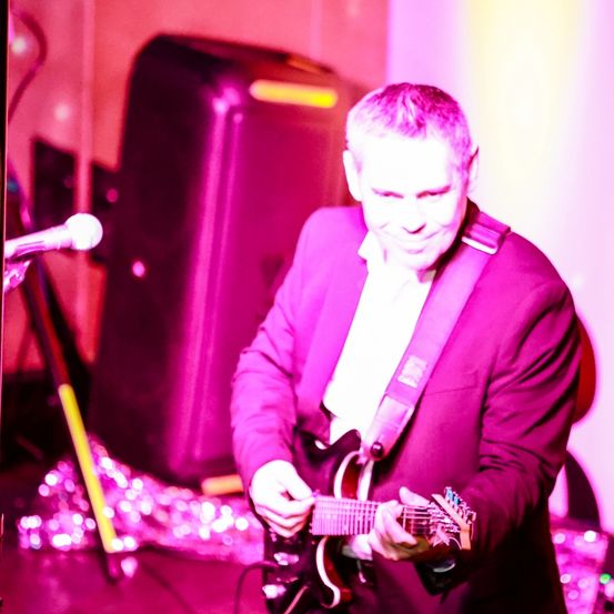 Ein Mann in einem lila Anzug spielt eine E-Gitarre unter einem rosa Licht bei einem Konzert.