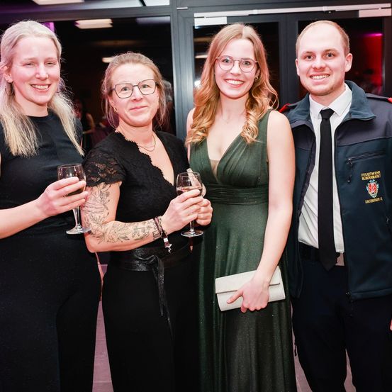 Vier Personen stehen eng beieinander, lächeln und halten Weingläser. Sie sind bei einer formellen Veranstaltung, und die Frau in der Mitte trägt ein grünes Kleid. Der Mann rechts ist in Uniform.