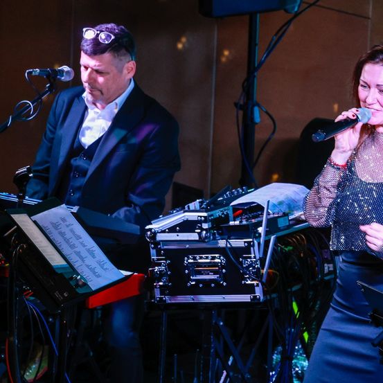 Ein Mann in Anzug spielt Klavier, während eine Frau in ein Mikrofon singt, beide von Bühnenlichtern beleuchtet.