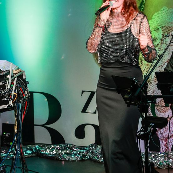 Eine Frau steht auf der Bühne und singt in ein Mikrofon vor einem Hintergrund mit den Buchstaben 'Z' und 'R'. Sie trägt ein mit Pailletten besetztes Oberteil und eine schwarze Hose. Um sie herum steht Equipment, mit grüner und blauer Beleuchtung.