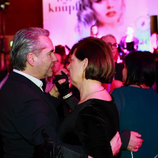 Ein Mann und eine Frau tanzen auf einer Party, beide in Schwarz gekleidet. Hinter ihnen zeigt eine große Leinwand das Gesicht einer Frau. Der Raum ist mit lila Lichtern schwach beleuchtet.