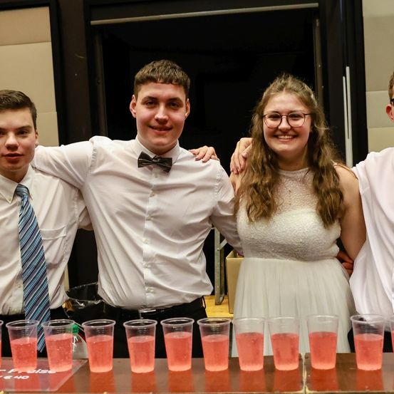 Vier junge Menschen stehen hinter einem Tisch mit Getränken davor. Der Mann in der Mitte trägt eine Fliege. Die Frau trägt eine Brille und ein weißes Kleid.