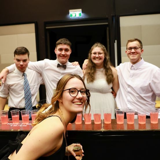Eine Gruppe junger Menschen lächelt und posiert für ein Foto an einer Bar. Es gibt rosa Getränke in Gläsern auf dem Tresen.