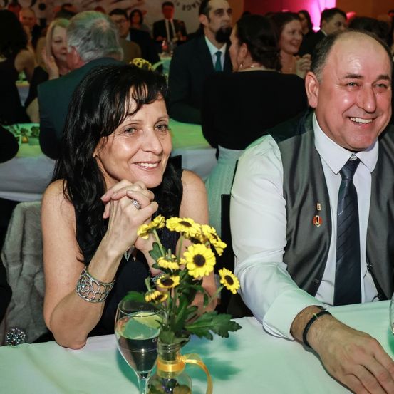 Ein Mann und eine Frau sitzen lächelnd an einem Tisch, mit einem Strauß Sonnenblumen im Vordergrund. Hinter ihnen sitzen andere Personen an Tischen bei einer Veranstaltung.
