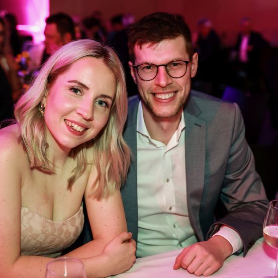 Ein Mann und eine Frau lächeln und posieren für ein Foto bei einer Veranstaltung. Sie sitzen an einem Tisch mit Weingläsern, und Menschen sind im Hintergrund verschwommen.