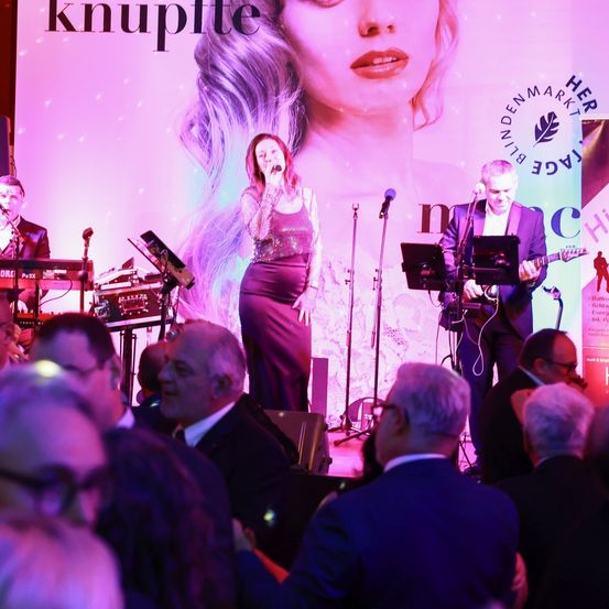 Eine Frau singt auf der Bühne mit einer Band vor einem großen Publikum. Die Bühne ist mit einem rosa Hintergrund und einem großen Poster dekoriert. Das Publikum sitzt und beobachtet die Aufführung.