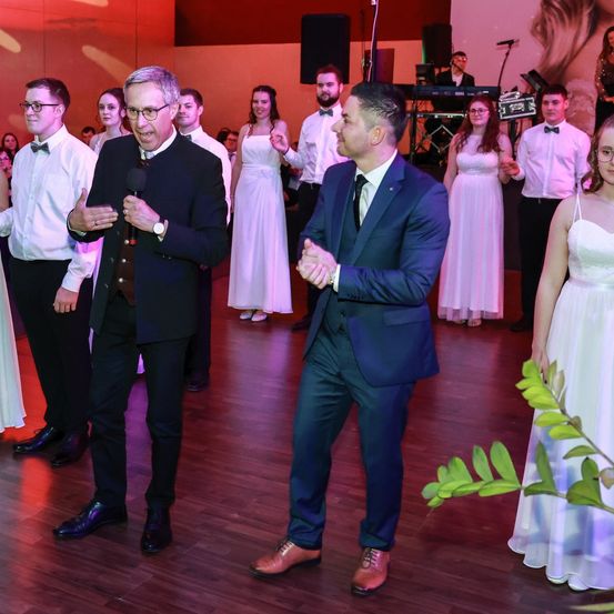 Eine formelle Veranstaltung mit Menschen in formeller Kleidung, ein Mann mit einem Mikrofon spricht, ein anderer Mann vor ihm mit einem Lächeln, eine Frau in einem weißen Kleid und eine grüne Pflanze auf dem Boden.