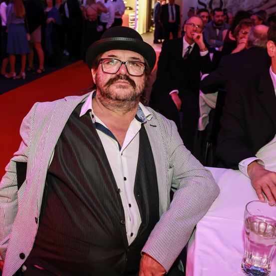 Ein Mann in Anzug und Hut sitzt bei einer formellen Veranstaltung an einem Tisch, vor sich ein Glas Wasser, mit mehreren Personen im Hintergrund.