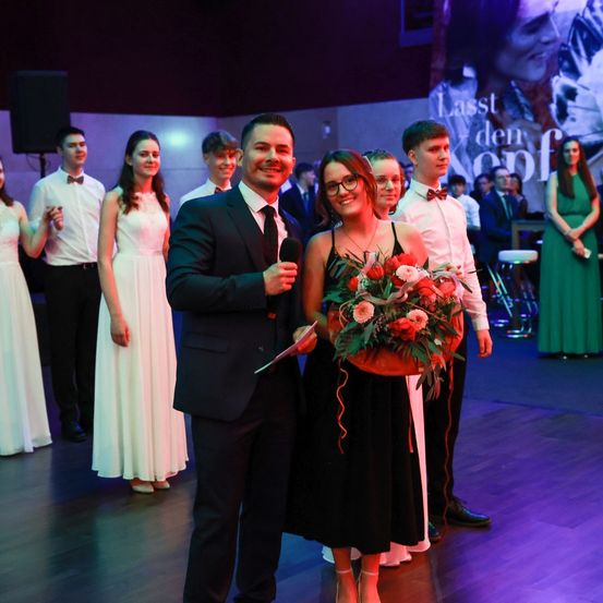 Ein Mann und eine Frau stehen lächelnd auf einer Bühne und halten einen Blumenstrauß. Hinter ihnen stehen mehrere Menschen in formeller Kleidung. Der Mann hält ein Mikrofon und die Frau trägt eine Brille. Im Hintergrund ist eine große Leinwand zu sehen.