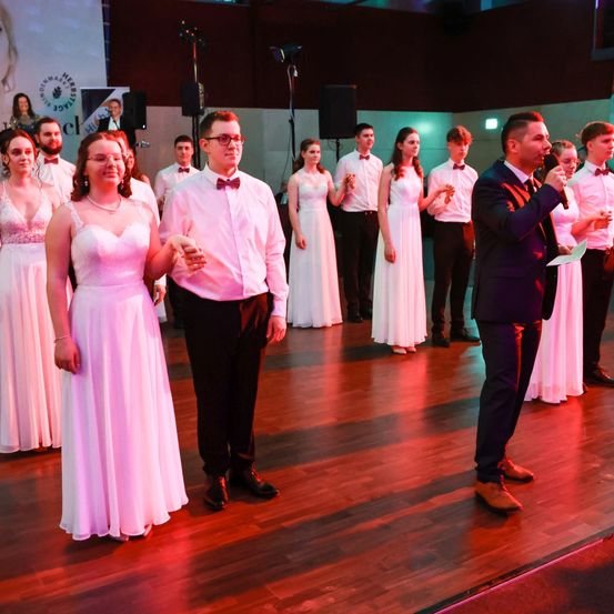 Eine Hochzeitsfeier mit einem Paar auf der Tanzfläche, einem Mann, der in ein Mikrofon spricht, und einer Gruppe von Menschen dahinter, einige klatschen.