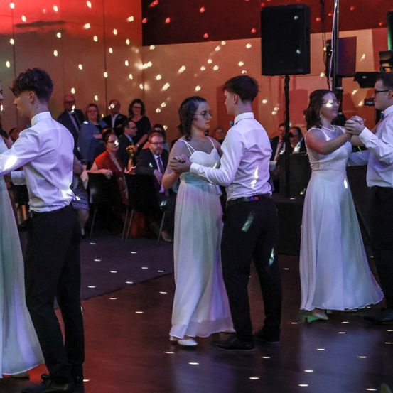 Mehrere Paare tanzen in einem Ballsaal mit roter Beleuchtung und Scheinwerfern, während andere von den Seiten zusehen. Der Raum ist mit Gästen gefüllt, einige sitzen und andere stehen.