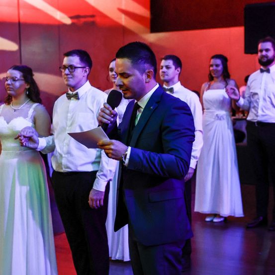 Ein Bräutigam hält eine Rede während einer Hochzeit und hält ein Papier und ein Mikrofon in der Hand. Die Braut und der Bräutigam stehen Hand in Hand und andere Gäste schauen zu.