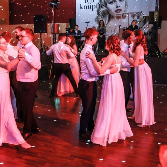 Eine Gruppe von Menschen in rosa Kleidern und Anzügen tanzt auf einem hölzernen Tanzboden. Eine große Leinwand zeigt eine Frau und einen Mann. Der Raum ist mit roten Lichtern beleuchtet.