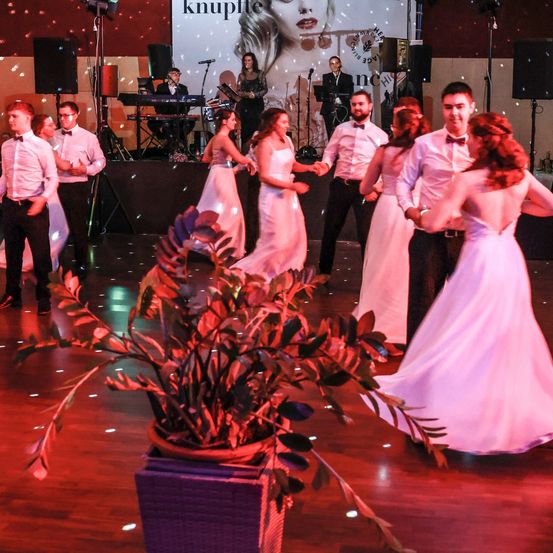 Eine Hochzeitsfeier mit einer Band auf der Bühne, bei der eine Frau in einem rosa Kleid mit einem Mann in Anzug und Fliege tanzt, während andere zusehen und ein Topf im Vordergrund steht.