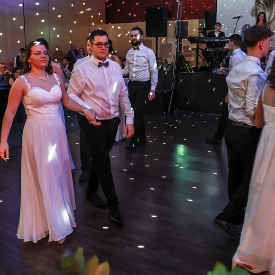 Ein Hochzeitsempfang mit einer von Diskolichtern beleuchteten Tanzfläche. Ein Paar tanzt in der Mitte, während andere am Rande zusehen. Im Hintergrund befindet sich eine Bühne mit einem Pianisten und einem Mikrofon.