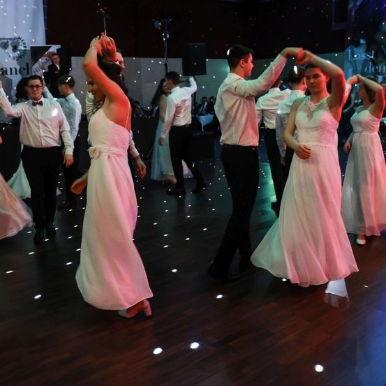 Eine Gruppe von Menschen tanzt auf einem Holzboden, gekleidet in formelle Kleidung. Die Frauen tragen rosa Kleider und die Männer weiße Langarmhemden und schwarze Hosen. Sie werden von Disco-Lichtern beleuchtet.