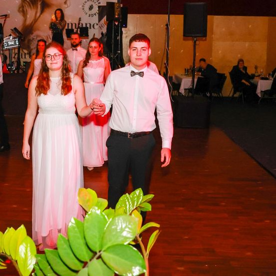 Ein Paar in pinker Kleidung tanzt auf einem Holzboden, während eine Band hinter ihnen spielt. Andere Personen sitzen an Tischen.