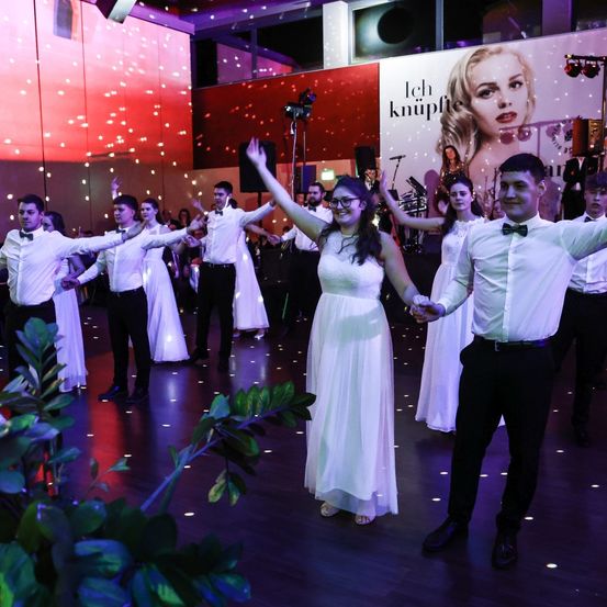 Eine Gruppe feierlich gekleideter junger Erwachsener tanzt auf einer Tanzfläche mit Disco-Lichtern und einer großen Werbung im Hintergrund.