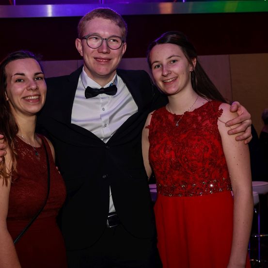 Drei Menschen posieren für ein Foto, zwei Frauen in roten Kleidern und ein Mann in einem schwarzen Anzug mit Fliege. Sie lächeln und stehen eng beieinander. Der Raum ist schwach beleuchtet.