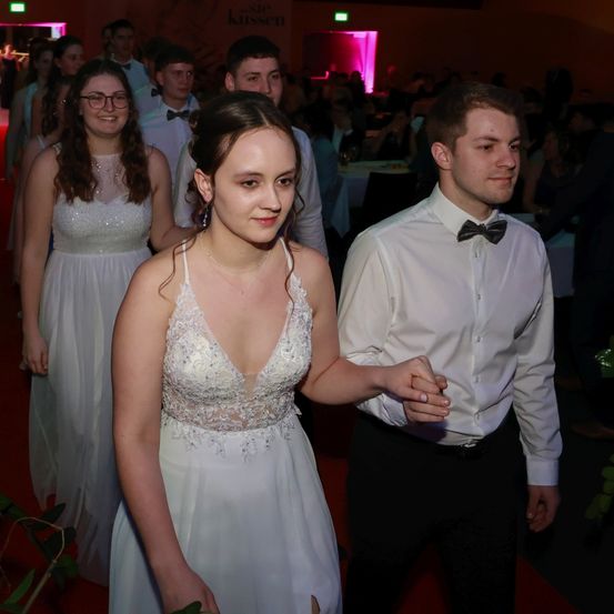 Eine junge Frau in einem weißen Kleid läuft mit einem Mann in formeller Kleidung den roten Teppich hinunter und beide lächeln in die Kamera. Hinter ihnen gehen auch eine Gruppe von ähnlich gekleideten Personen. Der Hintergrund ist schwach beleuchtet mit pinker Beleuchtung.