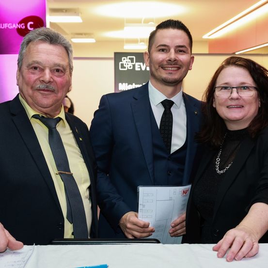 Drei Personen, zwei Männer und eine Frau, stehen eng zusammen in einem Konferenzsaal. Der Mann links trägt ein gelbes Hemd, der mittlere Mann einen marineblauen Anzug und die Frau eine Brille und ein schwarzes Oberteil. Sie lächeln und halten ein Dokument.