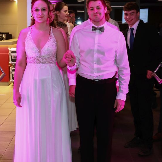 Eine Frau in einem weißen Kleid hält die Hand eines Mannes in einem rosa Hemd und Fliege. Sie stehen zusammen mit einem anderen Mann in einem schwarzen Anzug und blauer Krawatte.