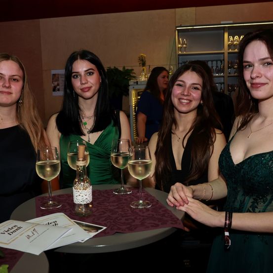 Vier Frauen sitzen um einen Tisch mit Weingläsern, eine hält eine Flasche. Sie tragen grüne Kleider, und Papiere mit 'Vielchen Dank' liegen auf dem Tisch. Eine Frau in einem blauen Kleid steht im Hintergrund in der Nähe eines Regals mit Gegenständen.