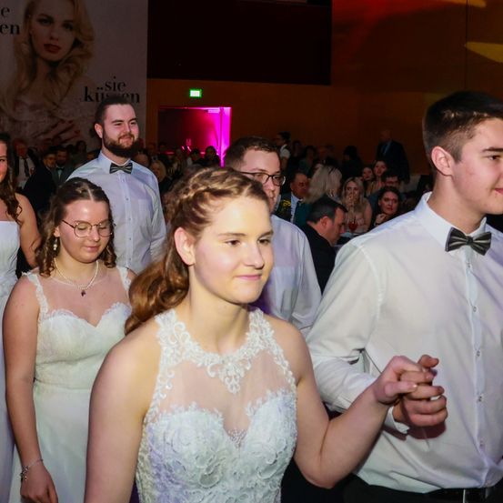 Eine Gruppe von Menschen ist in einem Saal versammelt. Eine Frau in einem weißen Kleid lächelt und geht mit einem Mann in einem weißen Hemd und Fliege. Hinter ihnen stehen zwei Frauen in weißen Kleidern, von denen eine eine Brille trägt. An der Wand hängt ein großes Plakat.