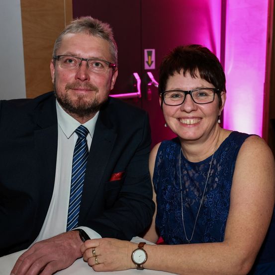 Ein Mann und eine Frau sitzen lächelnd zusammen an einem Tisch, mit einem rosa Licht hinter ihnen. Der Mann trägt einen dunklen Anzug und eine Krawatte, während die Frau ein blaues Kleid und eine Brille trägt.