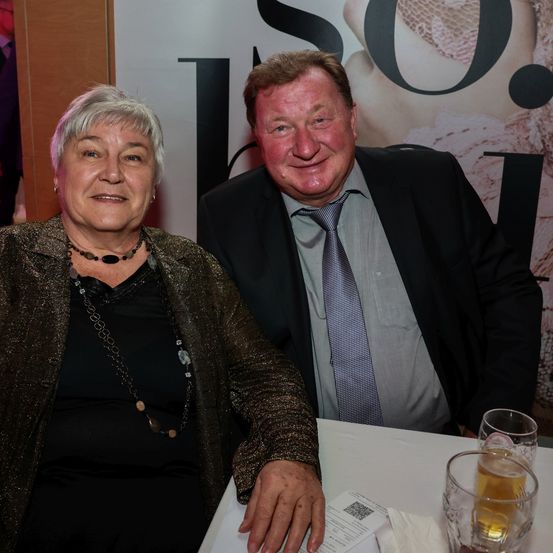 Eine ältere Frau und ein Mann lächeln in die Kamera. Sie sitzen an einem Tisch mit Getränken und scheinen sich bei einer Veranstaltung zu befinden.