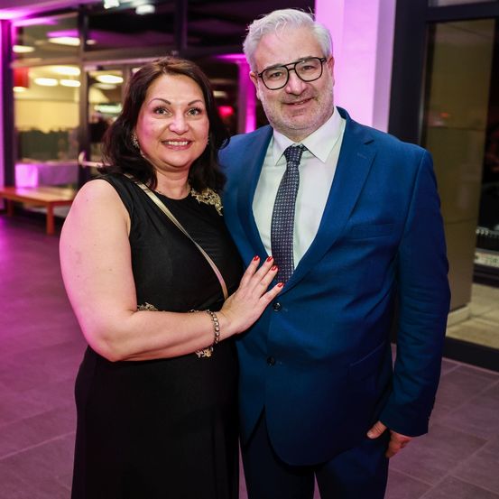 Ein lächelndes Paar steht bei einem Event zusammen, die Frau in einem schwarzen Kleid und der Mann in einem blauen Anzug mit gemusterter Krawatte, beide unter pinker Beleuchtung.