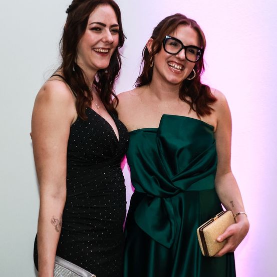 Zwei Frauen stehen lächelnd nebeneinander. Eine trägt ein schwarzes Kleid mit weißen Punkten, die andere ein grünes Kleid mit einer Schleife. Sie halten Handtaschen und stehen vor einer rosa Wand.