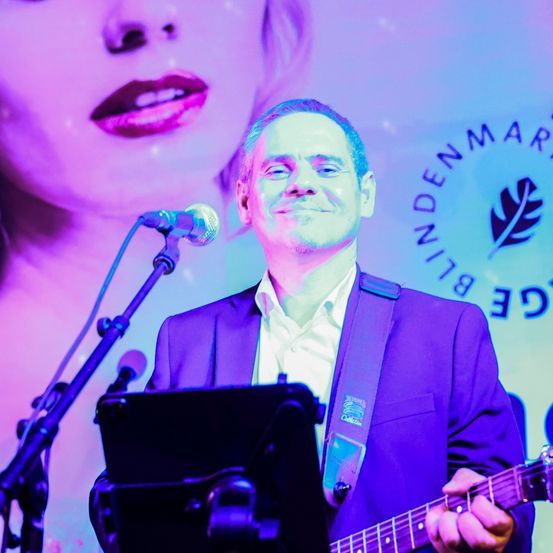 Ein Mann spielt Gitarre auf der Bühne, mit einem Mikrofon vor ihm. Dahinter ist ein großer Hintergrund mit dem Gesicht einer Frau und dem Logo 'BINDENDMARKET'.