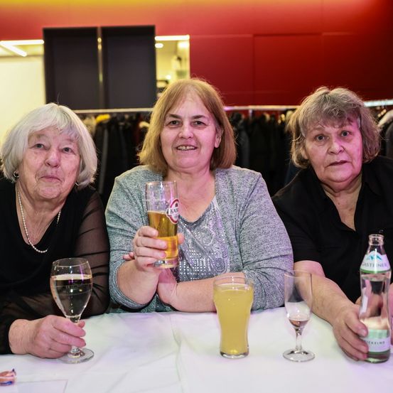 Drei ältere Frauen sitzen an einem Tisch, jede hält ein Glas mit einem Getränk. Die Frau in der Mitte lächelt und hält ein Glas Bier. Hinter ihnen hängen Kleidungsstücke an Kleiderbügeln.