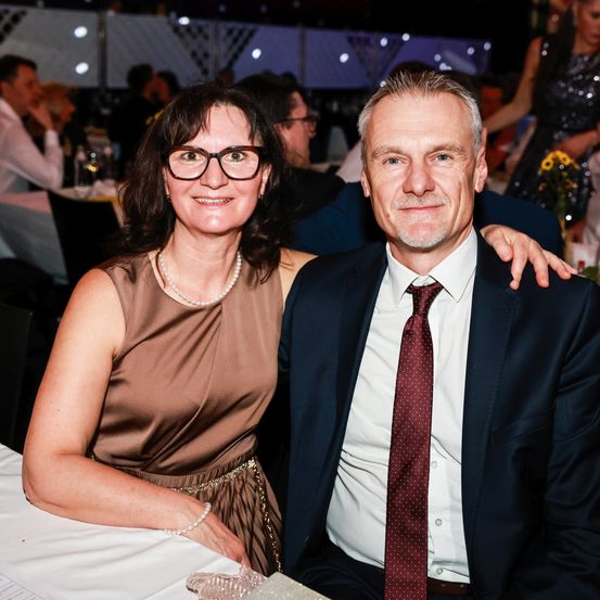 Ein Mann und eine Frau sitzen bei einer formellen Veranstaltung zusammen und lächeln. Die Frau trägt ein braunes Kleid und eine Brille, während der Mann eine rote Krawatte und einen Anzug trägt. Sie sind von einem unscharfen Hintergrund anderer Veranstaltungsteilnehmer umgeben.