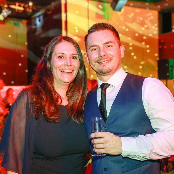 Ein Mann und eine Frau lächeln und posieren für ein Foto auf einer Party. Der Mann hält ein Glas Wein.