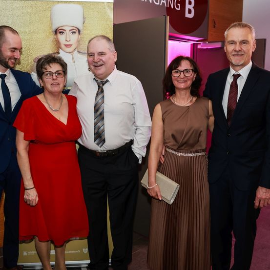 Fünf formell gekleidete Personen posieren für ein Foto vor einem Hintergrund mit einer Frau in Uniform.