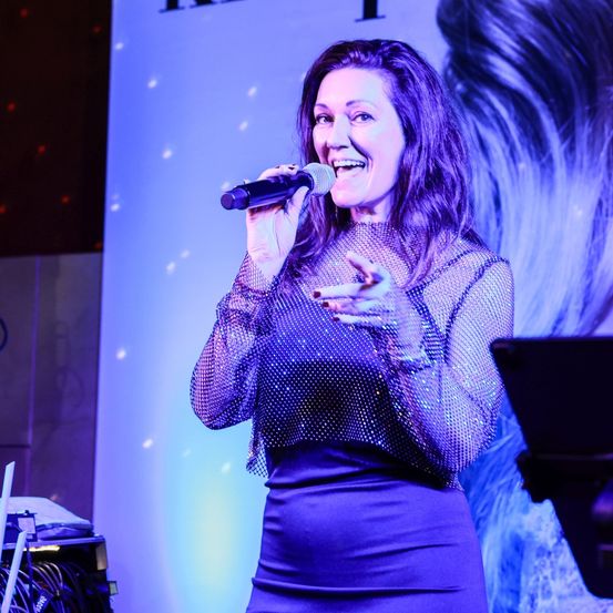 Eine Frau in einem lila Paillettenoberteil und -kleid singt in ein Mikrofon auf der Bühne mit lila Beleuchtung.
