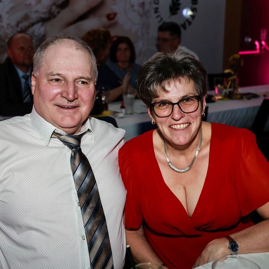 Ein Mann und eine Frau lächeln in die Kamera. Der Mann trägt eine Krawatte, und die Frau hat eine Brille und eine Halskette. Hinter ihnen sitzen Leute an Tischen, und ein großes Bild ist an der Wand.