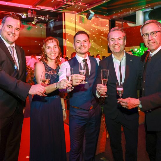 Fünf Menschen in formeller Kleidung lächeln und halten Weingläser in einer Partyumgebung. Der Hintergrund besteht aus einem roten Teppich und bunten Lichtern.