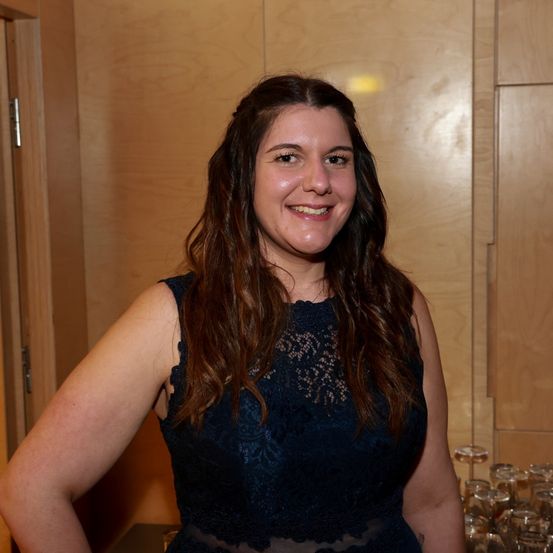 Eine Frau mit langen braunen Haaren lächelt in die Kamera. Sie steht drinnen und trägt ein blaues Spitzenkleid. Hinter ihr ist eine Holzwand.
