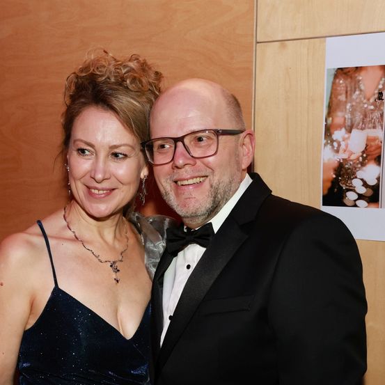 Ein lächelnder Mann und eine Frau in formeller Kleidung stehen zusammen in der Nähe einer Holzwand mit einem Bild einer Person in einem funkelnden Kleid.
