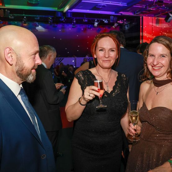 Drei Menschen lächeln bei einer formellen Veranstaltung. Eine Frau in der Mitte hält ein Glas Wein. Der Hintergrund zeigt eine bunte Umgebung mit roten und lila Lichtern.