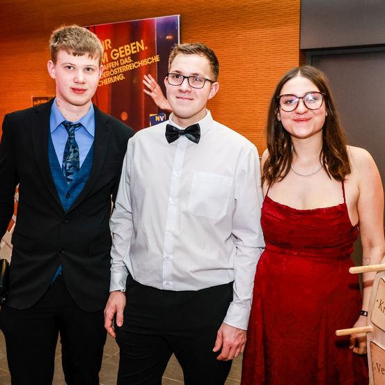 Drei junge Menschen stehen zusammen in einem Raum und posieren für ein Foto. Zwei sind Männer, eine ist eine Frau. Die Männer tragen Anzüge und Brillen, die Frau ein rotes Kleid. Ein Banner ist hinter ihnen sichtbar.