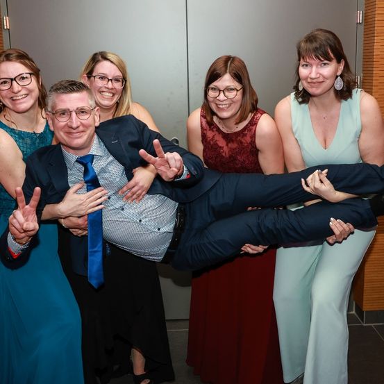 Eine Gruppe von Frauen in Kleidern posiert für ein Foto mit einem Mann im Anzug, alle lächeln. Der Mann in der Mitte macht ein Friedenszeichen. Die Frau auf der linken Seite trägt eine Brille. Der Raum hat eine graue Wand.