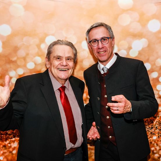 Zwei Männer stehen nebeneinander und lächeln für ein Foto. Sie sind in formeller Kleidung gekleidet. Der Mann auf der linken Seite trägt eine rote Krawatte.