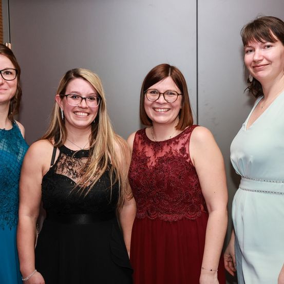 Fünf Frauen stehen lächelnd zusammen, wahrscheinlich bei einer Veranstaltung. Sie tragen verschiedene farbige Kleider und Brillen. Die Frau links trägt ein blaues Kleid, und die Frau neben ihr trägt ein schwarzes Kleid. Die Frau in der Mitte trägt ein rotes Kleid. Die Frau rechts trägt ein weißes Kleid.