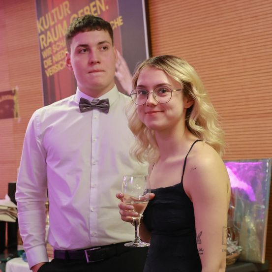 Ein Mann und eine Frau stehen lächelnd zusammen bei einer Veranstaltung. Der Mann trägt eine Fliege und hält ein Weinglas. Die Frau mit Tattoos trägt ein schwarzes Kleid und hat einen fröhlichen Gesichtsausdruck. Hinter ihnen ist eine Wand mit Werbetext.