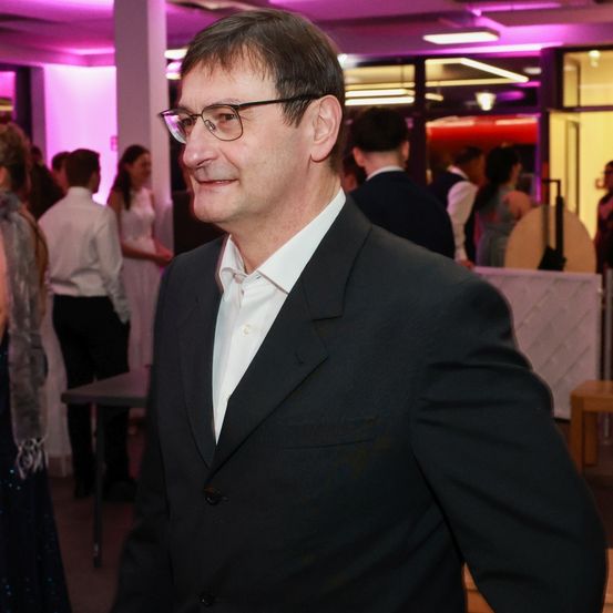 Ein Mann in einem schwarzen Anzug und mit Brille steht in einem Raum mit rosa Licht. Hinter ihm unterhalten sich mehrere Menschen. Links steht ein Tisch und rechts eine Holzbank.