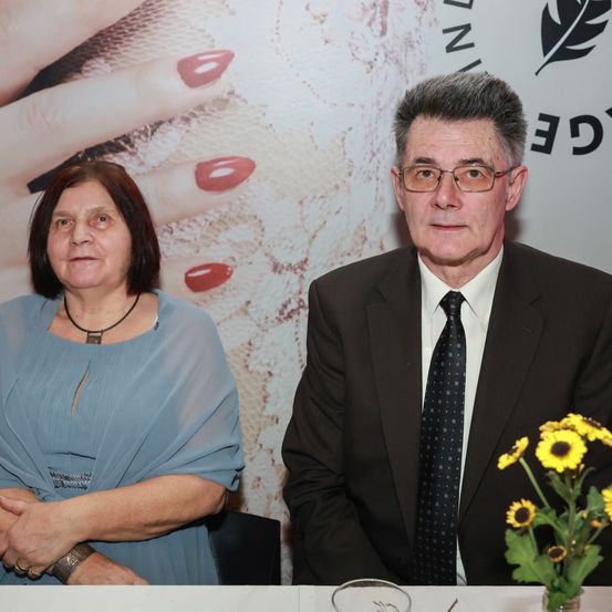 Eine ältere Frau und ein Mann sitzen an einem Tisch und posieren für ein Foto. Die Frau trägt ein blaues Kleid und der Mann einen schwarzen Anzug mit Krawatte. Ein Blumenvase steht auf dem Tisch. Hinter ihnen ist ein großes Bild von Händen mit roten Nägeln.